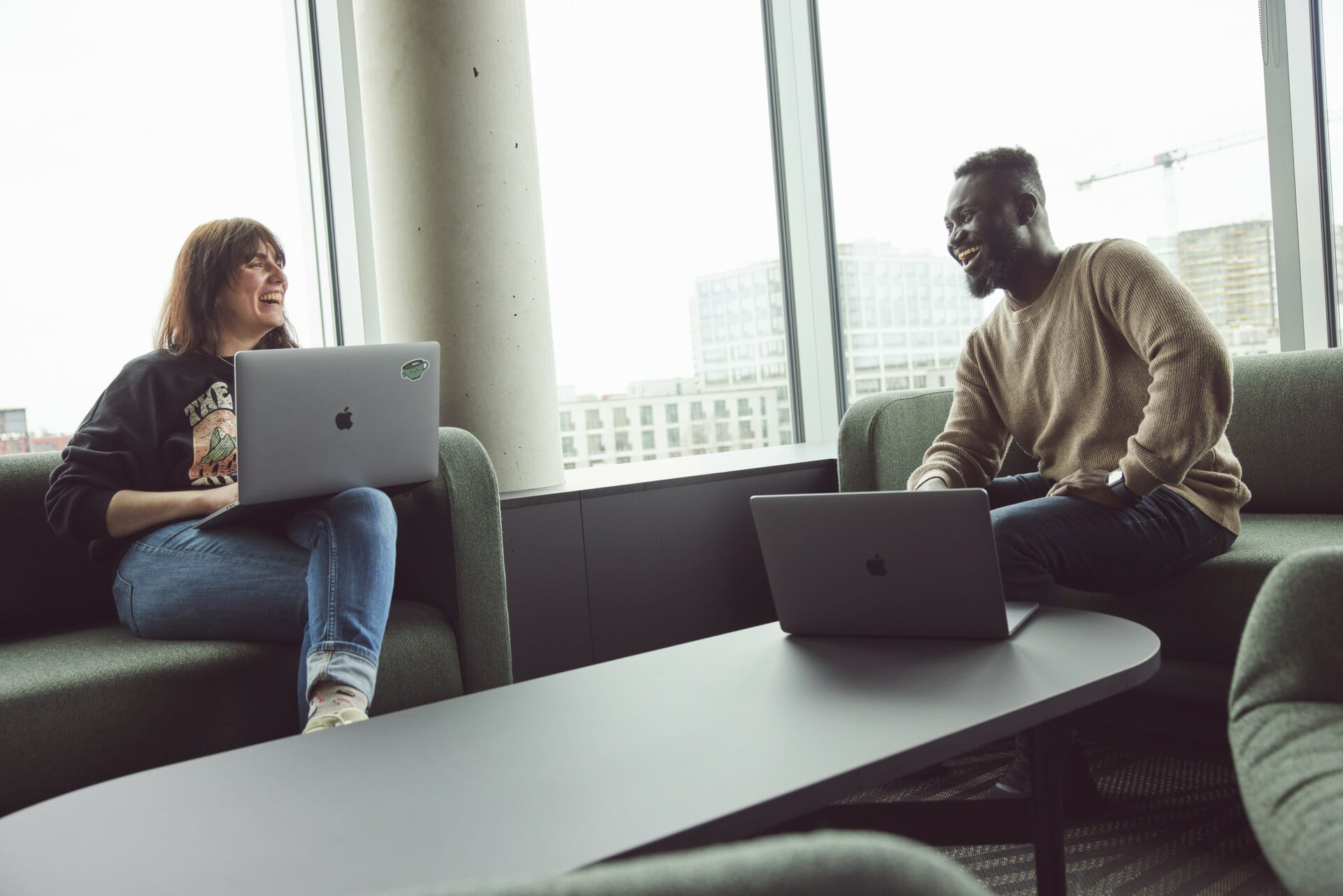 Two people sit on couches facing each other while working on laptops in a bright, modern office space with large windows, collaborating seamlessly as minubo partners.