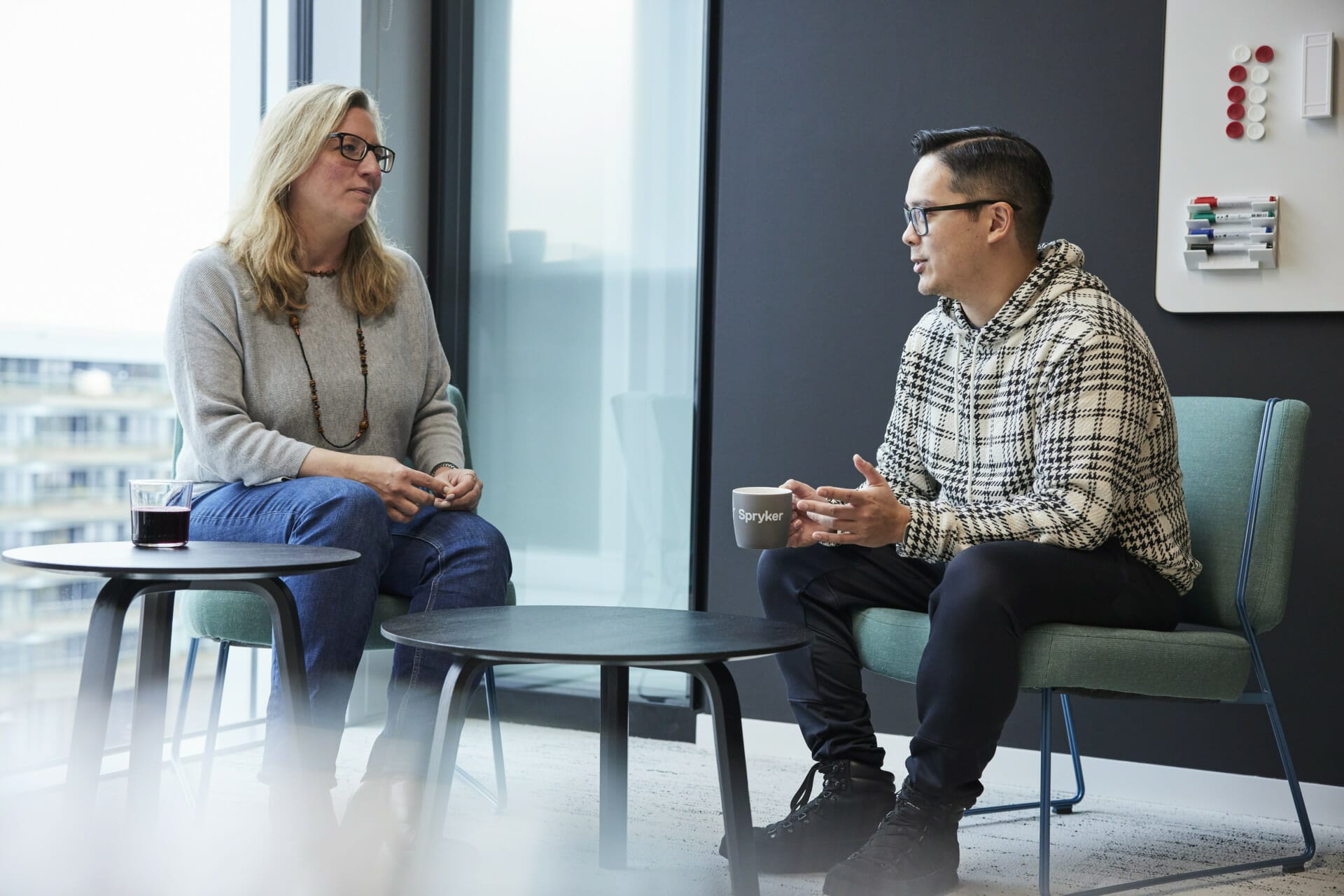 Two people sit and converse in a CriticalRiver office setting, one holding a coffee mug. They are seated on chairs around a low table, with a window in the background and a whiteboard on the wall.