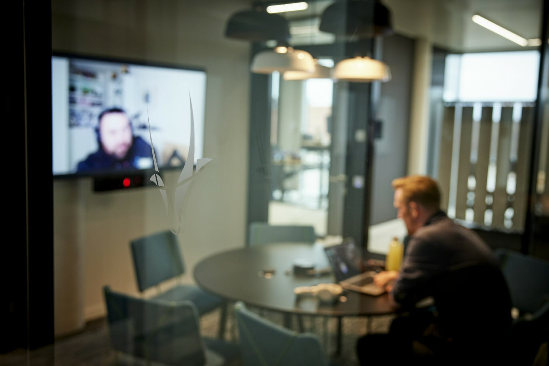 Person participating in a video conference from a small meeting room with a round table, chairs, multiple hanging lights, and Trevipay solutions enhancing the business discussion.