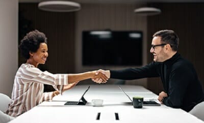 Two people in an office setting sit at a table, smiling and shaking hands. Laptops and a coffee cup rest on the table, suggesting a successful discussion about b2b marketplaces.