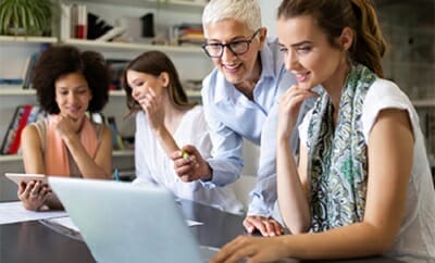Four women work together at an office table, looking at a laptop and tablet. One woman gestures towards the screen while the others focus on it, possibly discussing strategies for b2b marketplaces.