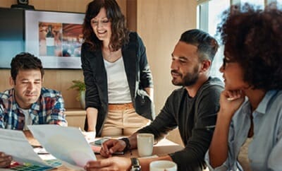 Four people are sitting around a table looking at documents, with one standing and smiling. Coffee cups and various papers cover the table, creating an atmosphere of focused collaboration, typical in b2b marketplaces.