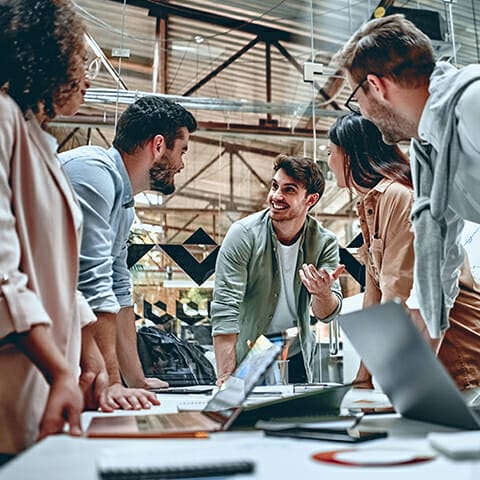 A group of five people are gathered around a table in a modern office, actively discussing b2b marketplaces, with one person gesturing while speaking. Laptops and documents are placed on the table.