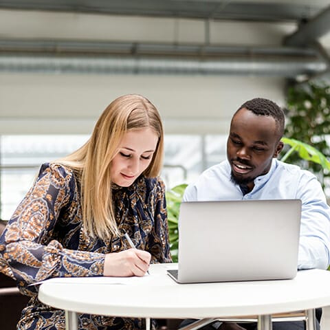 A woman and a man are working together at a round table. The woman is writing on a paper, while the man is looking at a laptop, possibly exploring b2b marketplaces. They are in a brightly lit room with large windows and plants in the background.