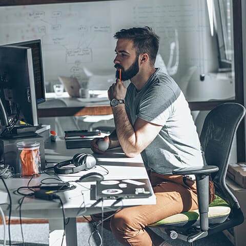 A man with a beard sits at a desk, focused on a computer screen. He holds an apple, with a pen in his other hand. The desk is cluttered with items like a laptop, headphones, and various knick-knacks—perhaps illustrating the busy world of composable commerce. A whiteboard stands in the background.