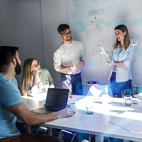A group of four people in a meeting room, engaged in discussion about composable commerce. They are standing or sitting around a table with laptops and papers. A whiteboard with diagrams is in the background.