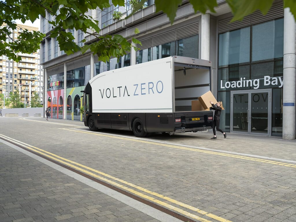 A person loads cardboard boxes into the back of a Volta Trucks' Zero electric delivery truck parked at a building's loading bay on a city street.