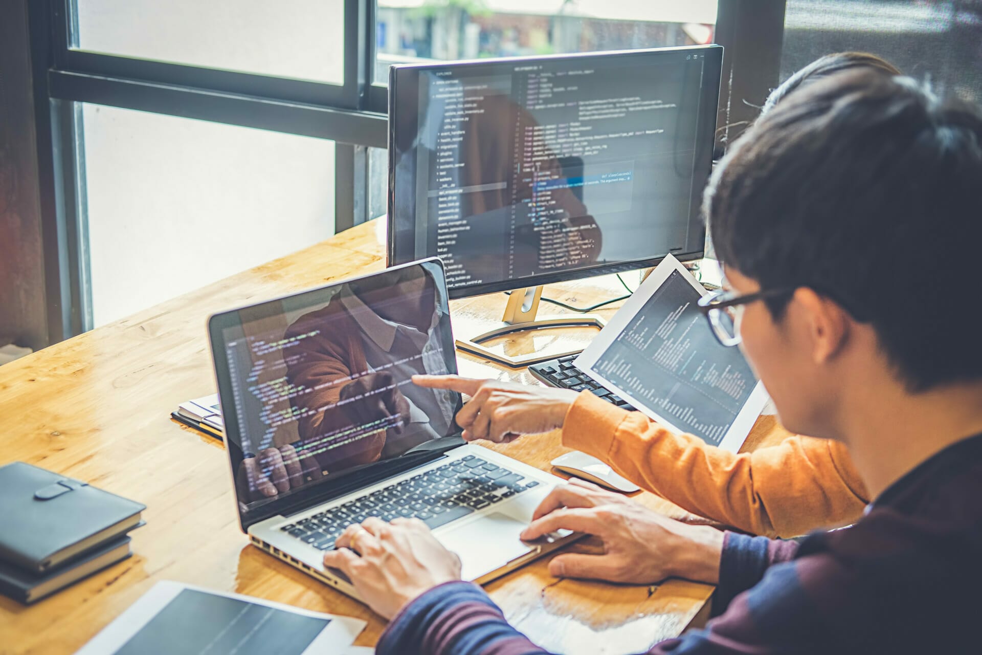 Two individuals work on greenfield software development using laptops and monitors, displaying code in a bright office setting. One points at the laptop screen while the other types. A tablet and notebook are on the desk.