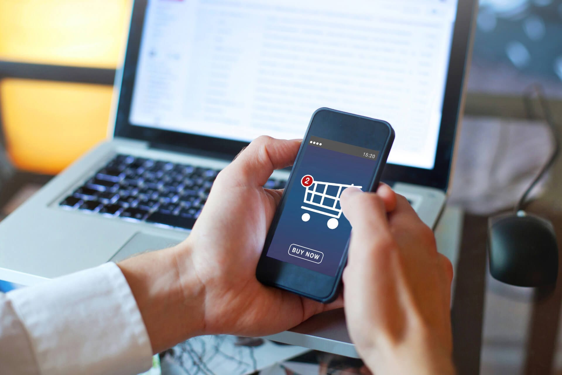 A person holds a smartphone displaying a shopping cart app with a "Buy Now" button and a notification showing two items. The phone is held in front of an open laptop, with the screen showing an email inbox. A computer mouse is placed beside the laptop.
