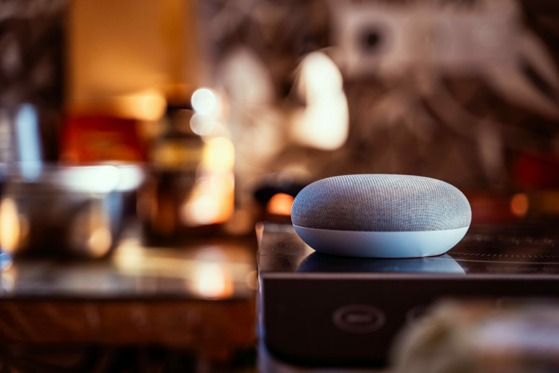 A smart speaker sits on a kitchen counter, subtly blending into its surroundings while facilitating seamless commerce through its IOT capabilities. Various out-of-focus kitchen items add to the scene's homely ambiance.