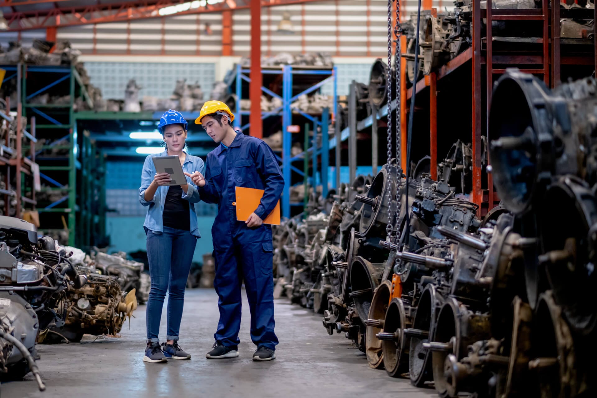 Two workers wearing safety helmets, one in blue and the other in yellow, stand in a large warehouse filled with machinery parts. They are looking at a tablet and discussing something. Shelves stocked with various metal components and engines are visible around them.