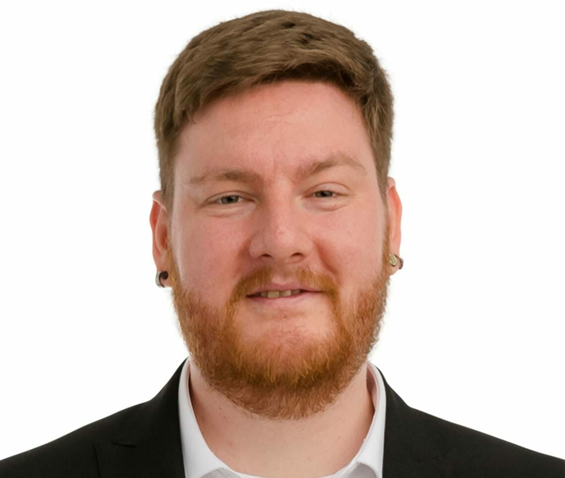 A man with short brown hair and a beard, wearing a black suit jacket, white shirt, and earrings, is smiling against a white background at Spryker EXCITE 2022.