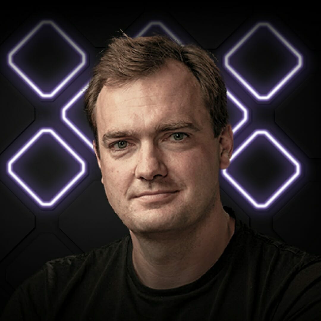 A man with short, light brown hair and a black shirt stands in front of a dark background illuminated by purple geometric neon shapes at EXCITE 2022 Nashville.