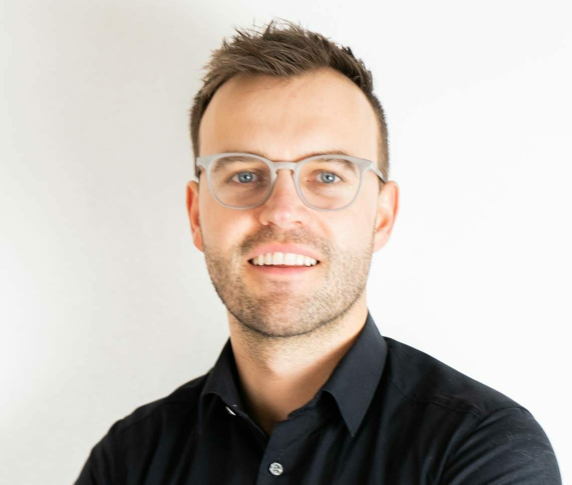 A man with short hair and glasses, wearing a black shirt, smiles while standing against a plain white background at the Spryker EXCITE 2022 event.