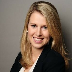 A woman with long blonde hair smiles while she wears a black blazer and white top, embodying the professionalism required for B2B Order Management, against a plain background.