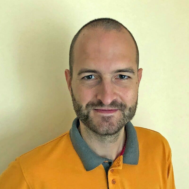 A bald man with a beard, representing the customer advisory board, is wearing an orange polo shirt with a grey collar and standing against a beige background.