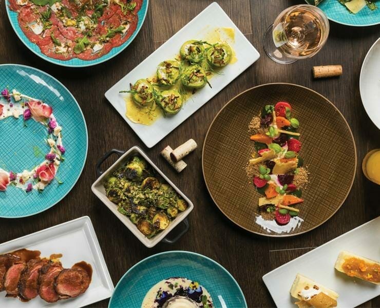 A variety of gourmet dishes, including meat, vegetables, and desserts, arranged on a dark wooden table with blue and brown plates, accompanied by glasses of drinks and wine corks—perfect for sparking some Shoptalk in Las Vegas.