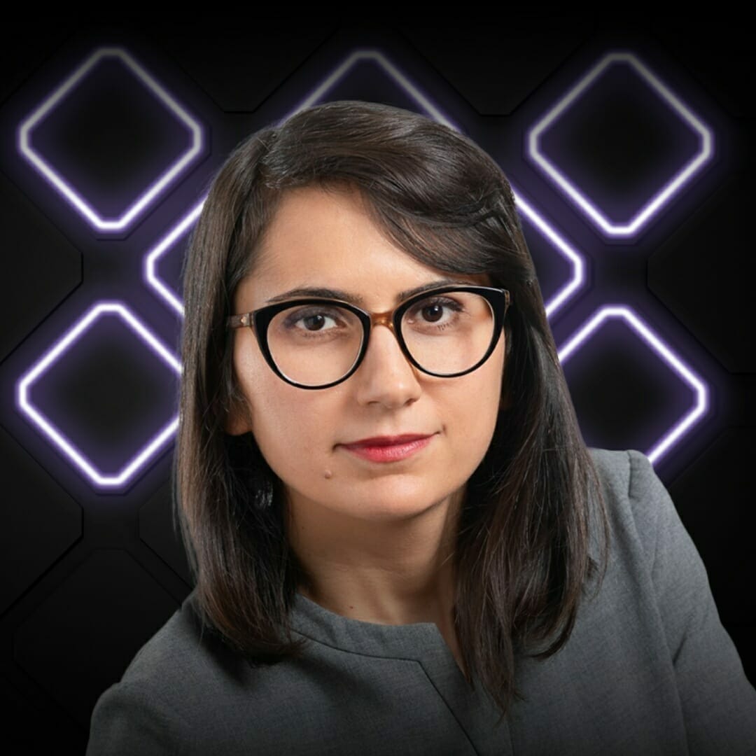 A woman with straight dark hair and glasses, wearing a gray outfit, poses in front of a dark background with glowing, square-shaped patterns at Spryker EXCITE 2022.