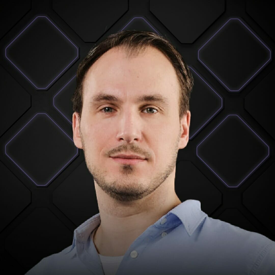 A man with short dark hair and a trimmed beard wears a light blue shirt at Spryker EXCITE 2022. The background consists of a black pattern with illuminated hexagonal shapes.