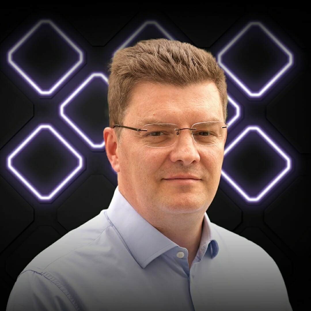 A man with glasses is wearing a light blue shirt, standing in front of a dark background with illuminated diamond patterns, capturing the vibrant energy of EXCITE 2022 Nashville.