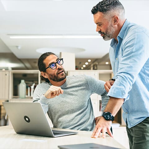 Two men talk, one is sitting next to a computer and the other is standing