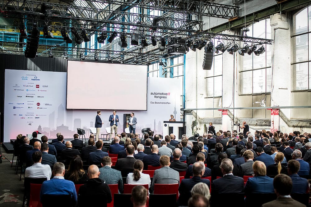 A conference room filled with seated attendees facing a stage where three presenters stand. The background includes large windows and a backdrop with logos from the automotiveIT Kongress. Event lighting and stage equipment are visible.