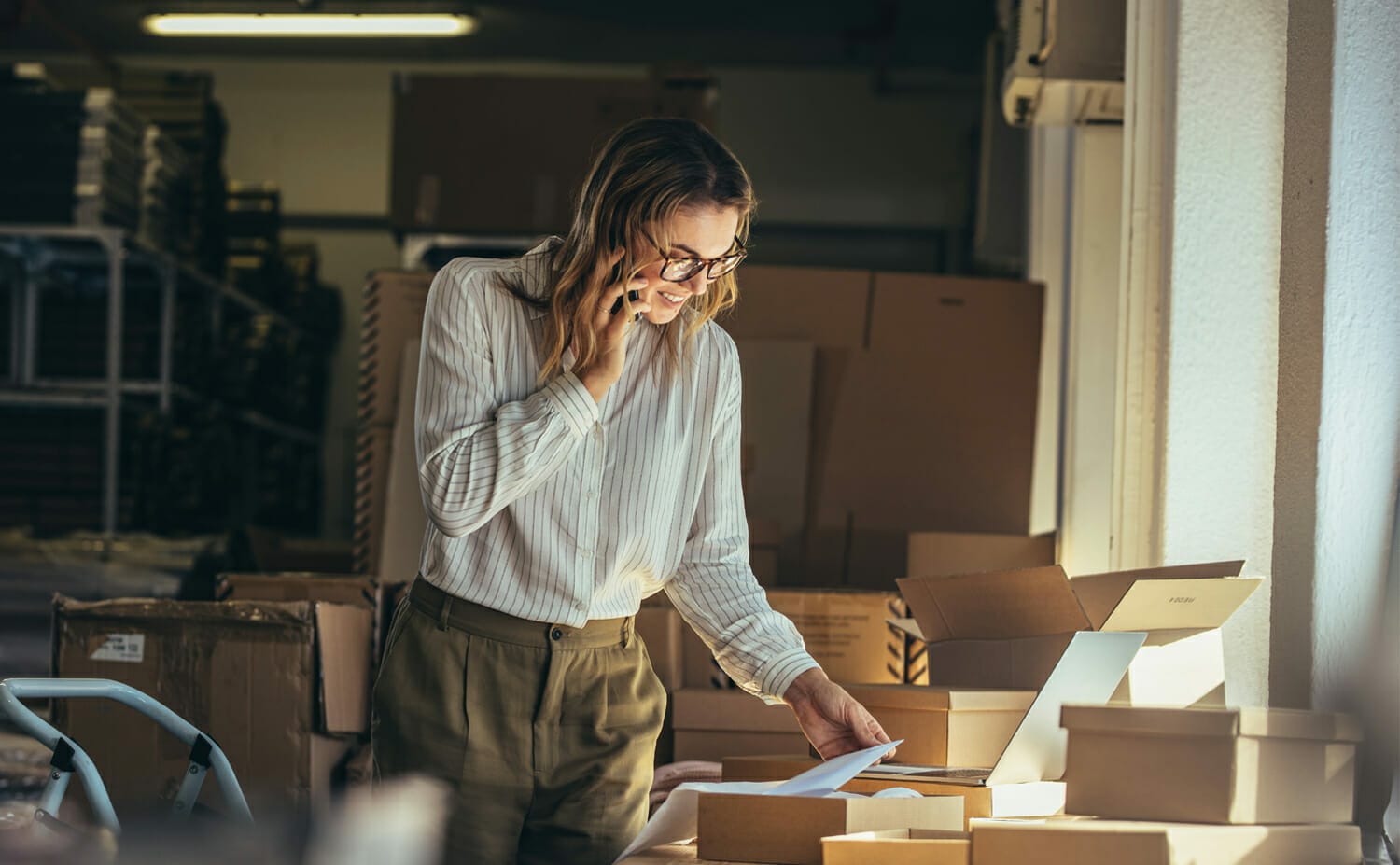 A person in a light-striped shirt and glasses is talking on the phone while standing next to a table with open cardboard boxes in what appears to be a warehouse, indicative of CPG commerce activities.