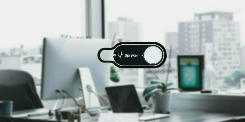 Close-up of a small black button labeled "Spryker" in an office setting with a computer, desk, and window overlooking buildings in the background. This Dash Button can be customized with SEO keywords to enhance your workspace efficiency.