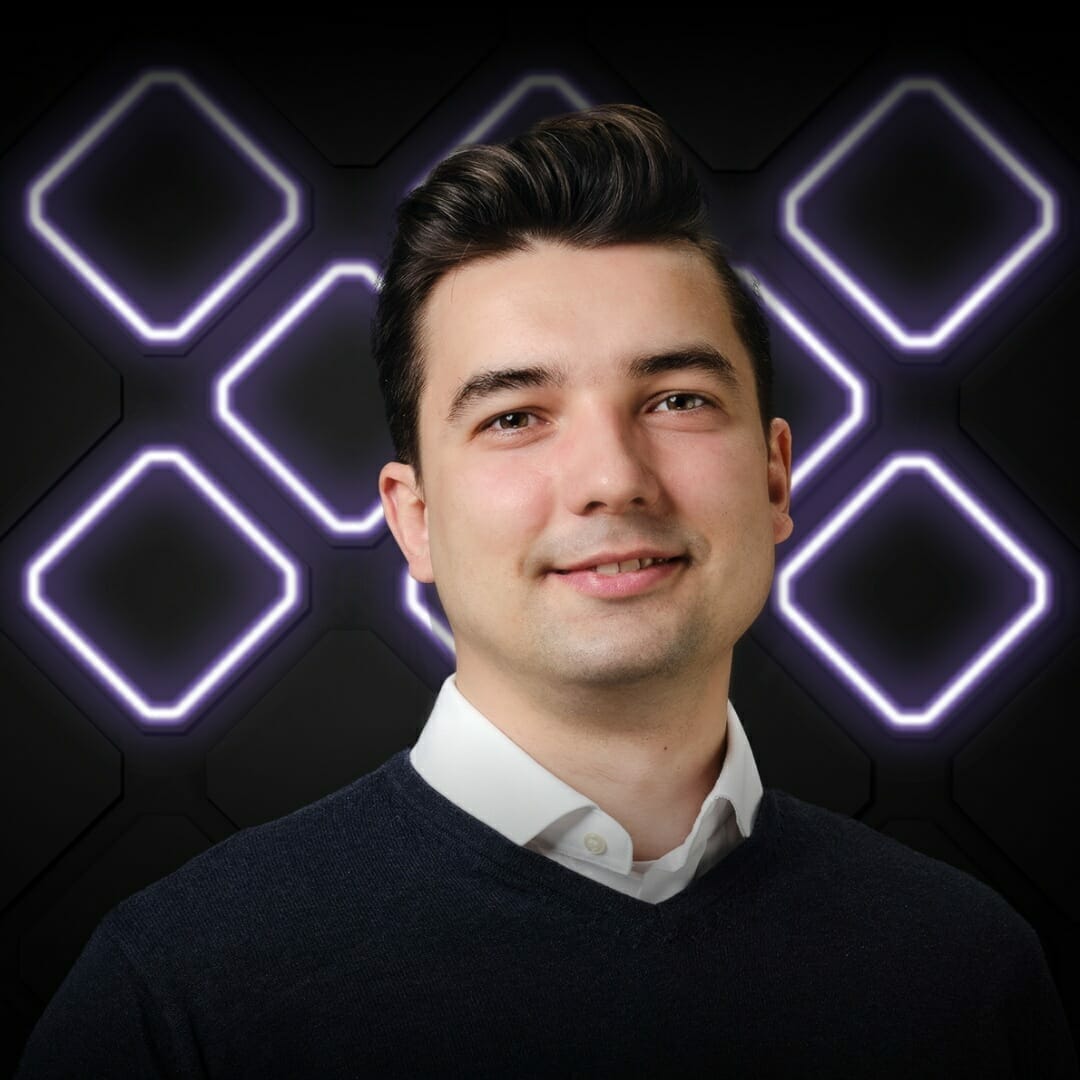 A person wearing a white shirt and a dark sweater smiles in front of a geometric, illuminated background with glowing squares at Spryker EXCITE 2022.