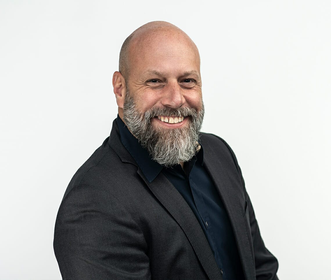 A bald man with a beard, smiling, wearing a dark blazer and shirt, poses confidently against a plain white background, embodying the professional spirit of Spryker at Shoptalk Europe.