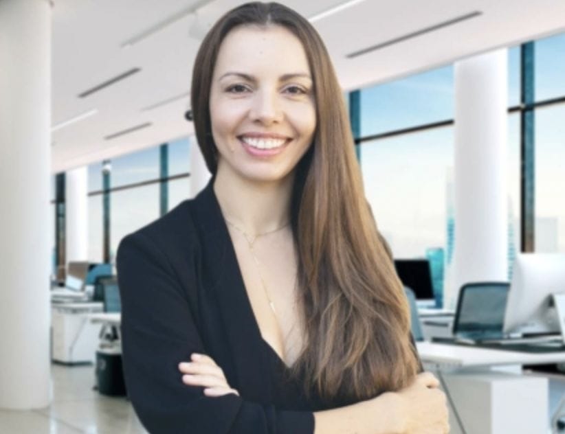 A person with long brown hair and a black blazer stands, arms crossed, in a bright, modern office with large windows. They exude confidence, ready to tackle the day's tasks and craft compelling press releases.