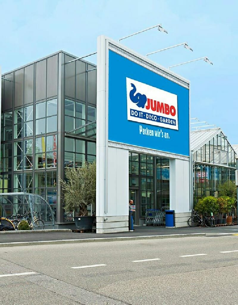 A store entrance with a large "Maximo JUMBO" sign, along with "Do It", "Deco", and "Garden". Several parked cars are visible in the foreground as if waiting for Marco Knecht himself to arrive.