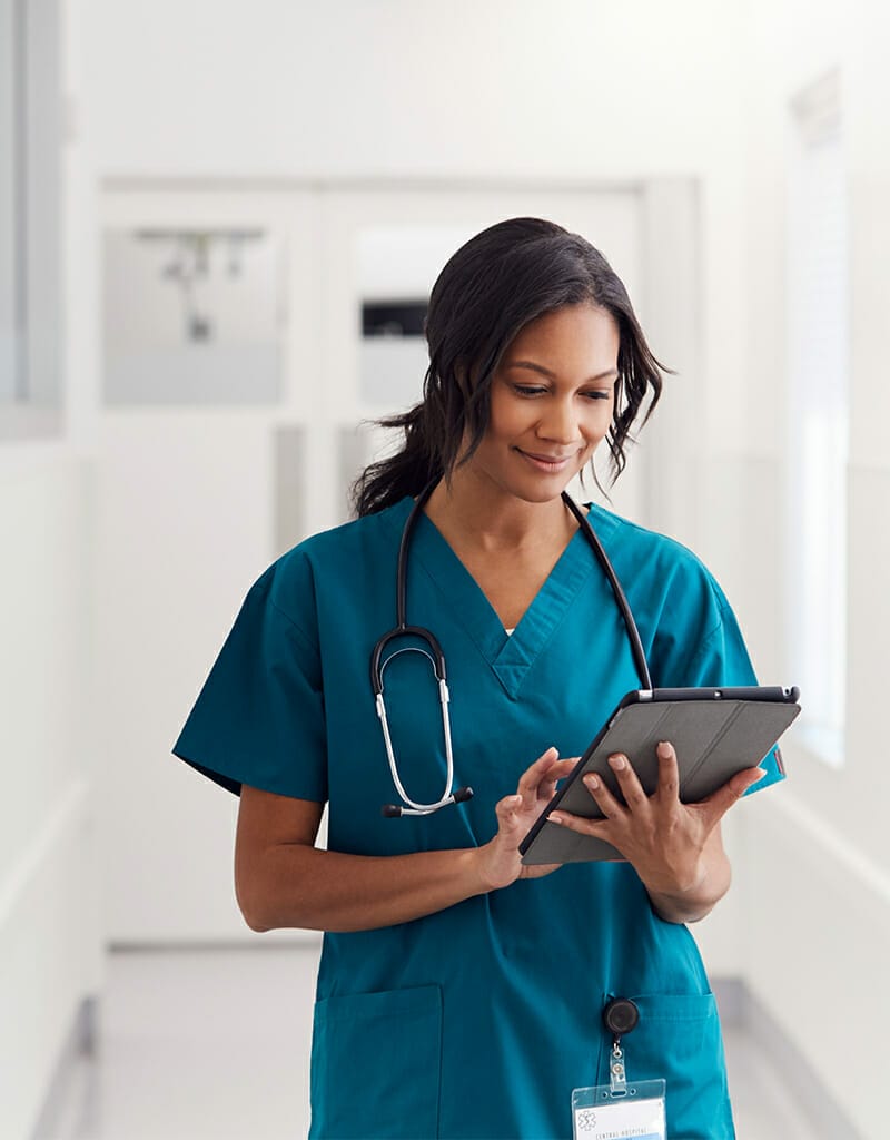 A medical professional in scrubs and a stethoscope around her neck looks at a tablet while standing in a well-lit hospital corridor, reminiscent of the efficiency showcased on Spryker OnAir.