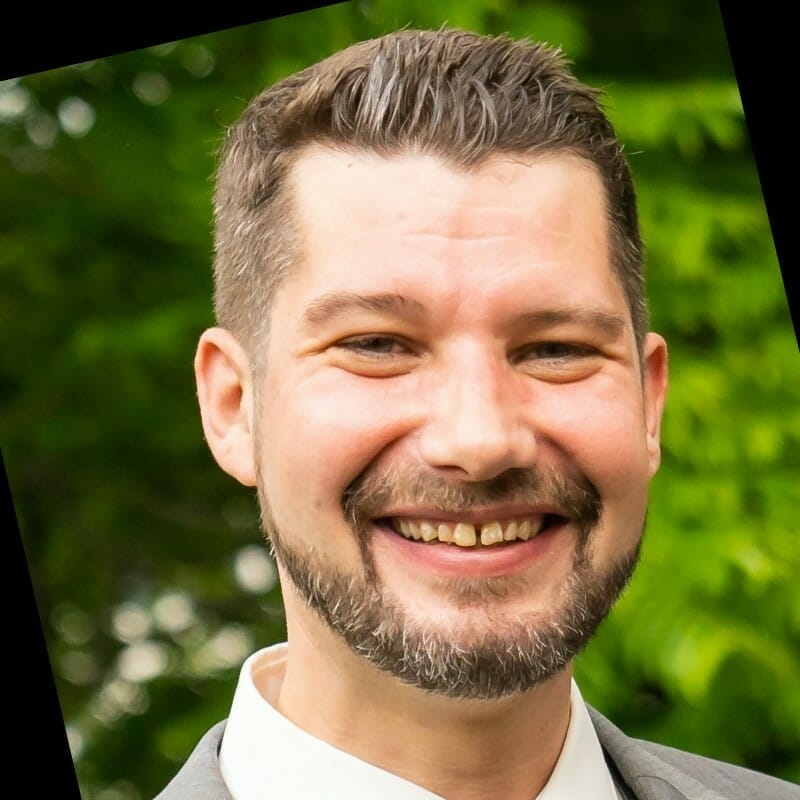 A man with a beard and mustache smiling, wearing a gray suit jacket and white shirt, stands in front of lush greenery. He’s ready for the customer advisory board meeting.