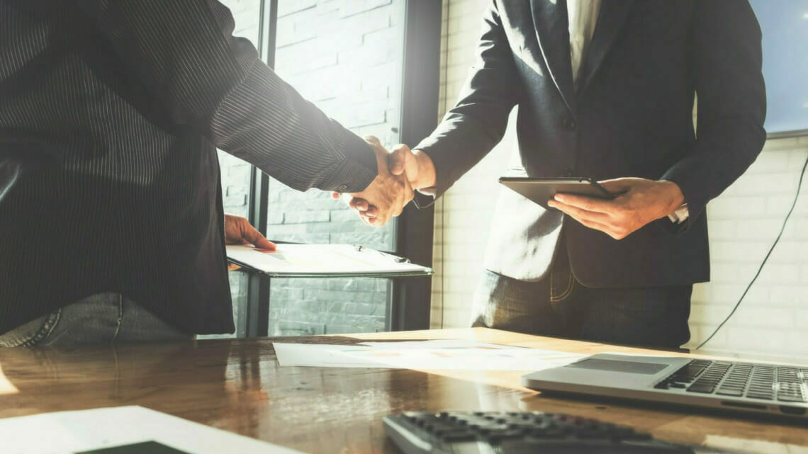 Two business professionals dressed in suits shake hands across a desk with documents and a laptop, indicating a business agreement. The scene captures the essence of an IDC Spryker collaboration meeting.