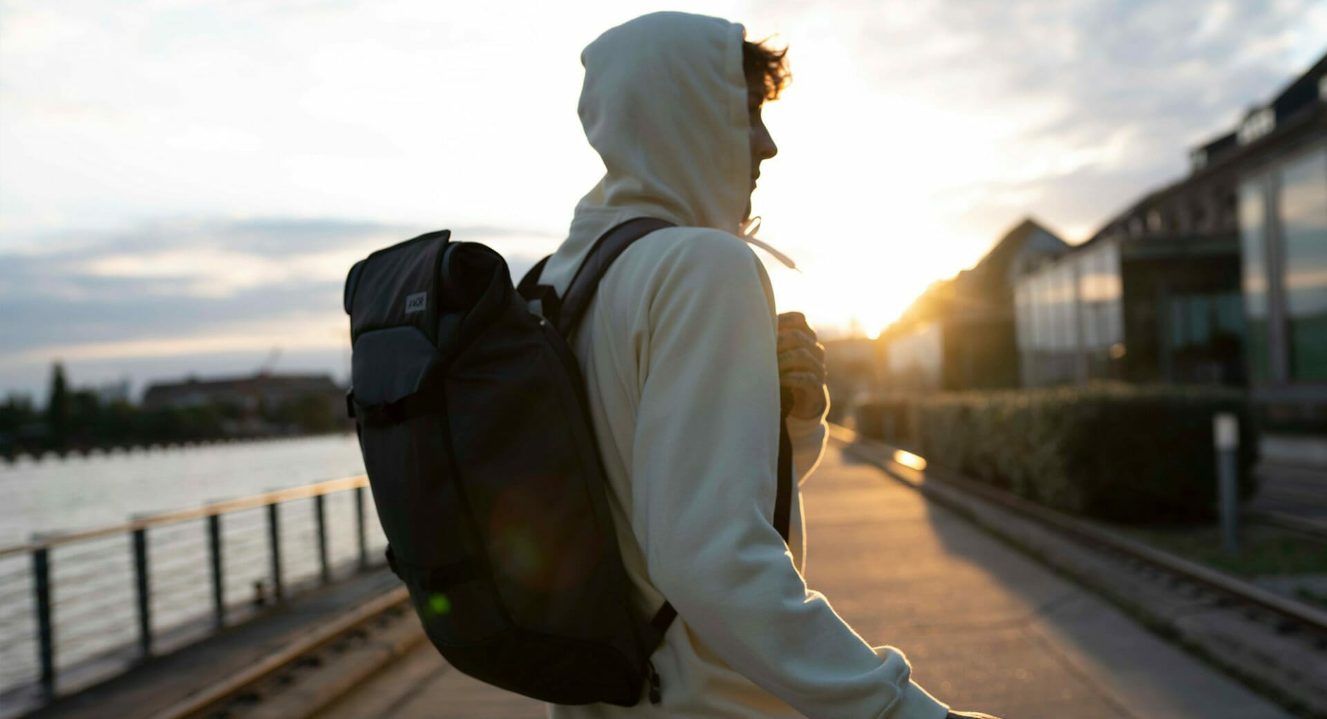 A person wearing a hoodie and carrying a backpack, fond of evening strolls, walks along a waterfront path at sunset.