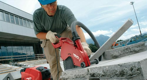 A construction worker wearing a blue helmet and safety gloves uses a red power saw to cut through concrete at a construction site, ensuring the precision and quality that spryker customers expect.