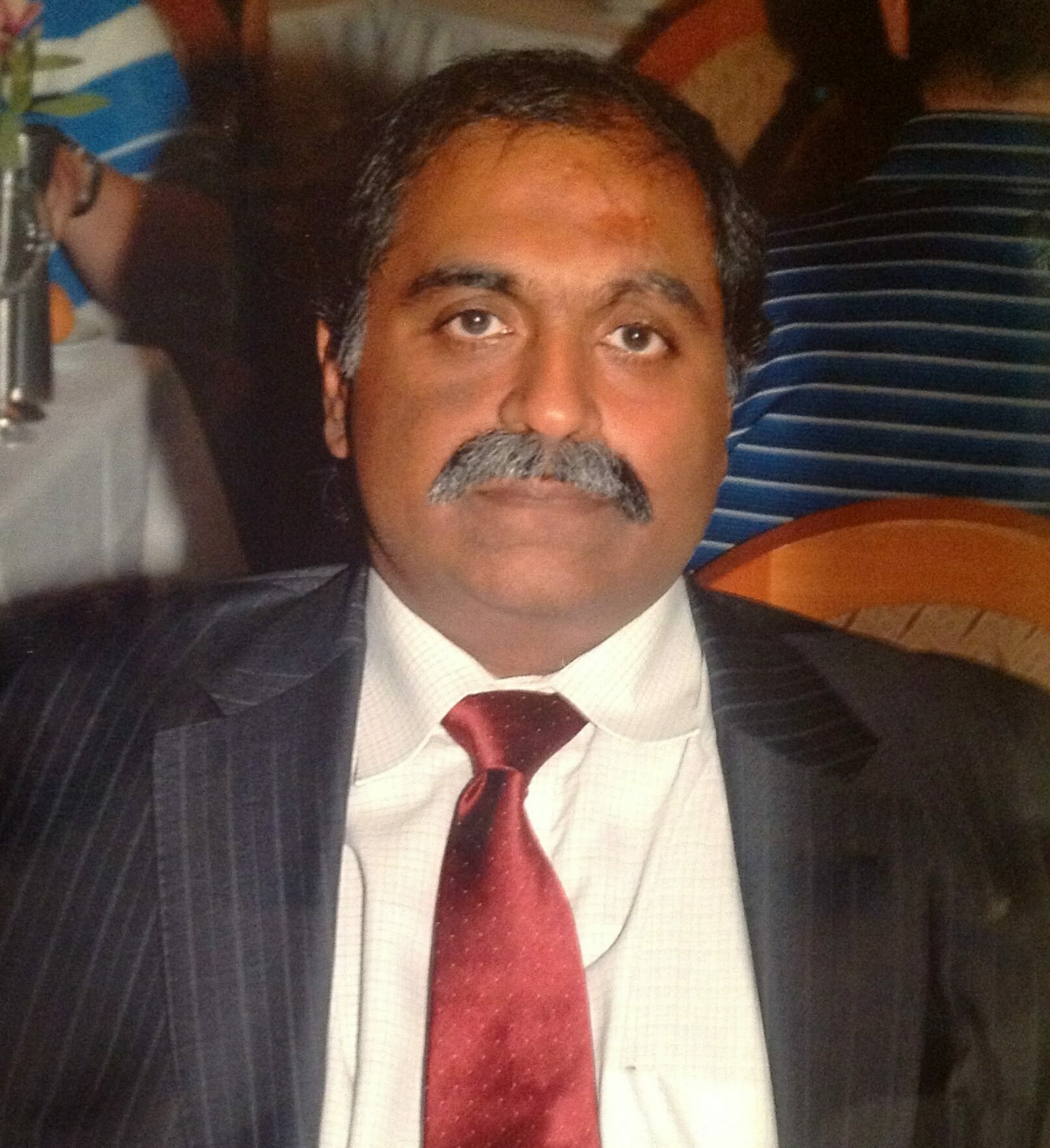A man with a mustache, wearing a dark suit, white shirt, and red tie, is seated at a table during a customer advisory board event.