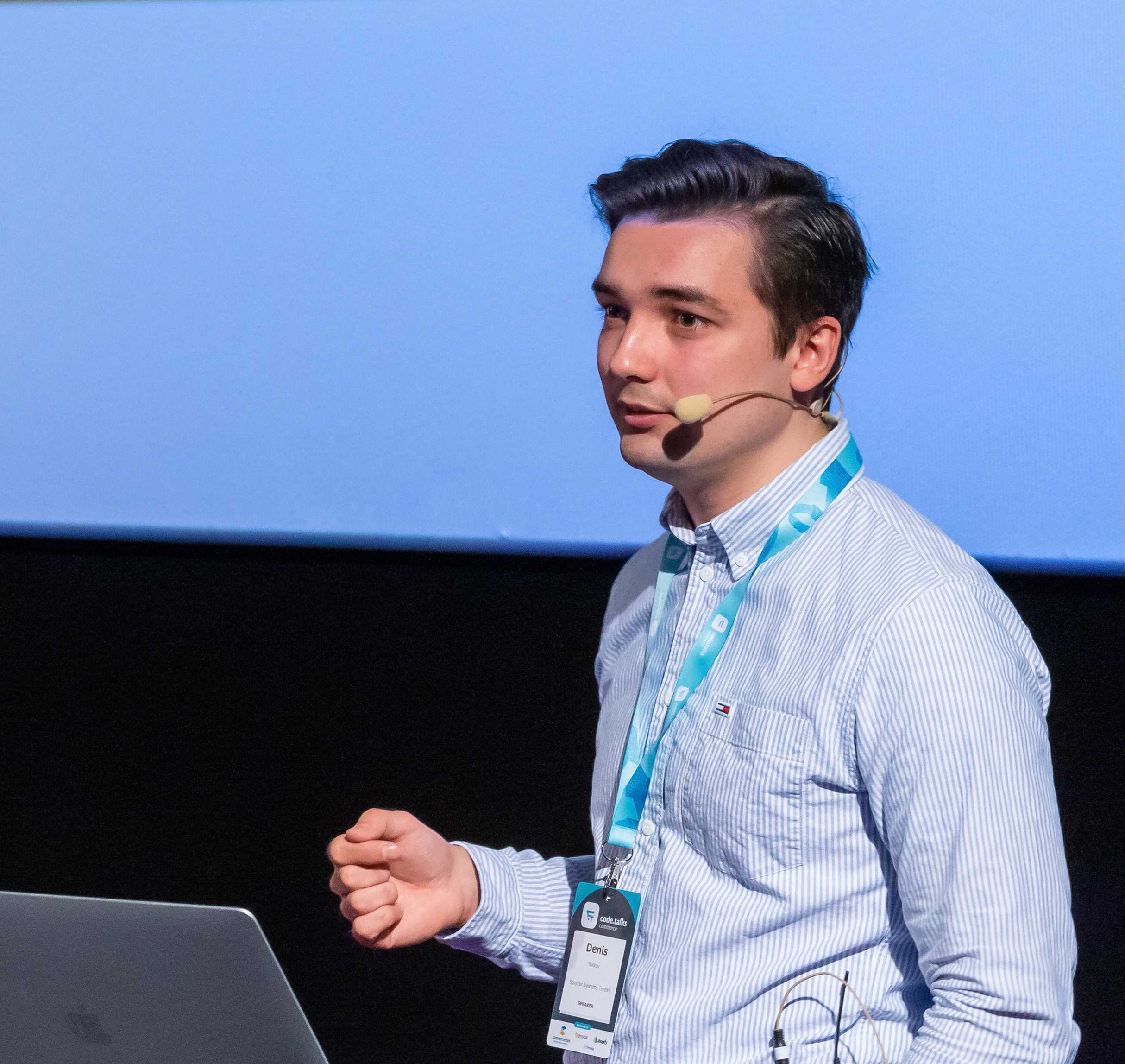 A person with a headset microphone is speaking on stage next to a laptop, wearing a striped shirt and an EXCITE conference name badge.