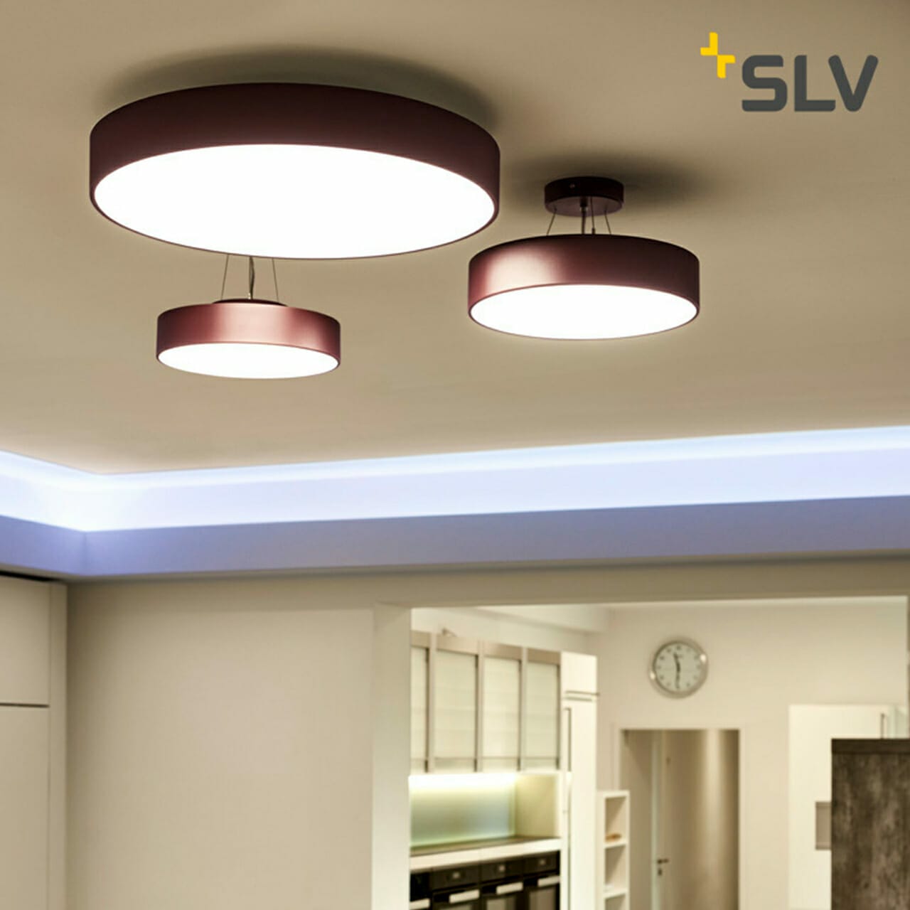 Three round ceiling lights with purple bases illuminate a modern, white kitchen featuring a clock on the wall. The lights are of different sizes, adding a touch of elegance to the space. The "SLV" logo in the upper right corner emphasizes their collaboration with a leading digital company.