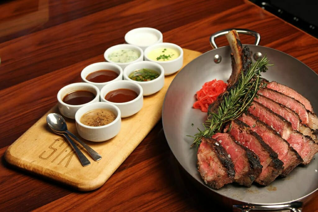 A sliced, medium-rare steak garnished with rosemary and pickled vegetables is served in a metal pan, accompanied by six small bowls of various sauces on a wooden board with spoons—perfect for some culinary shoptalk in Las Vegas.
