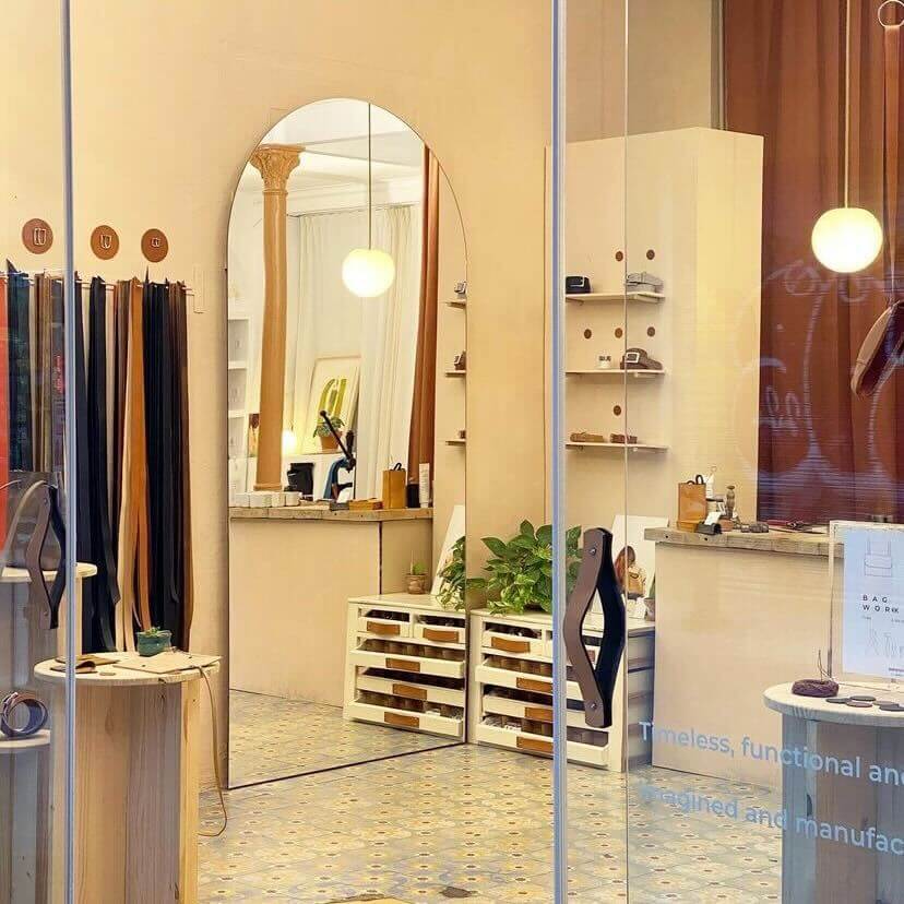A cozy, modern store interior with a large oval mirror, shelves displaying various items, potted plants, and neatly organized wooden crates. The decor includes hanging fabric swatches and a round ceiling light illuminating the space.