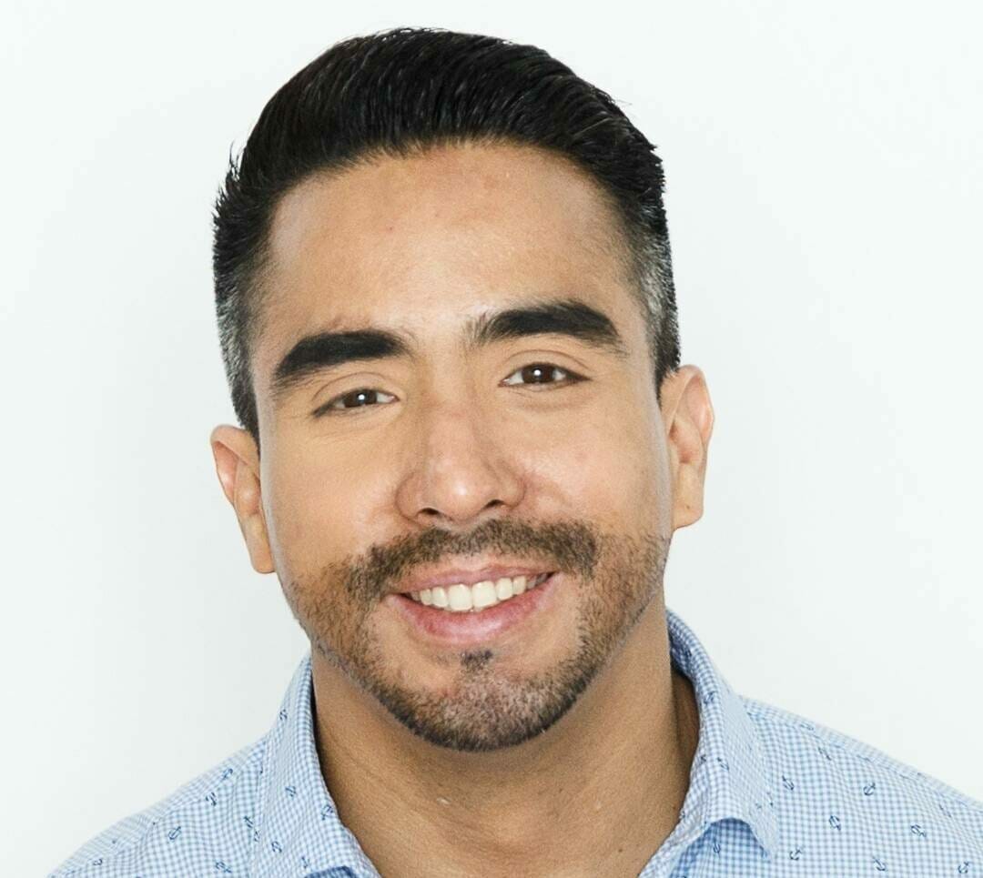 A man with short, dark hair and a trimmed beard smiles at the camera. He is wearing a light blue, button-up shirt with a subtle pattern. The background is plain and white, reminiscent of event portraits from Spryker EXCITE 2022.