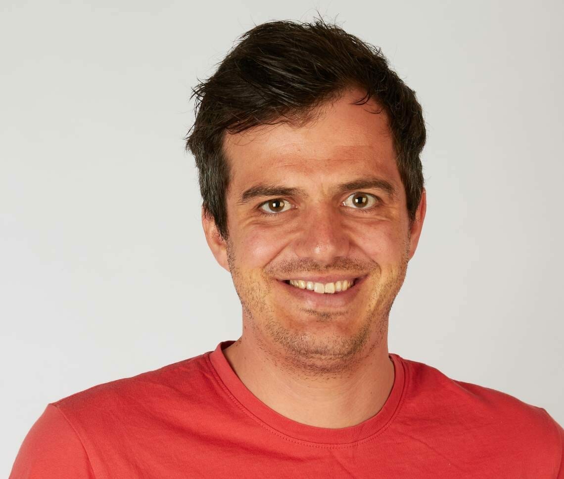 A man with short dark hair and a light beard is smiling, wearing a red shirt showcasing Spryker EXCITE 2022, and standing against a plain white background.