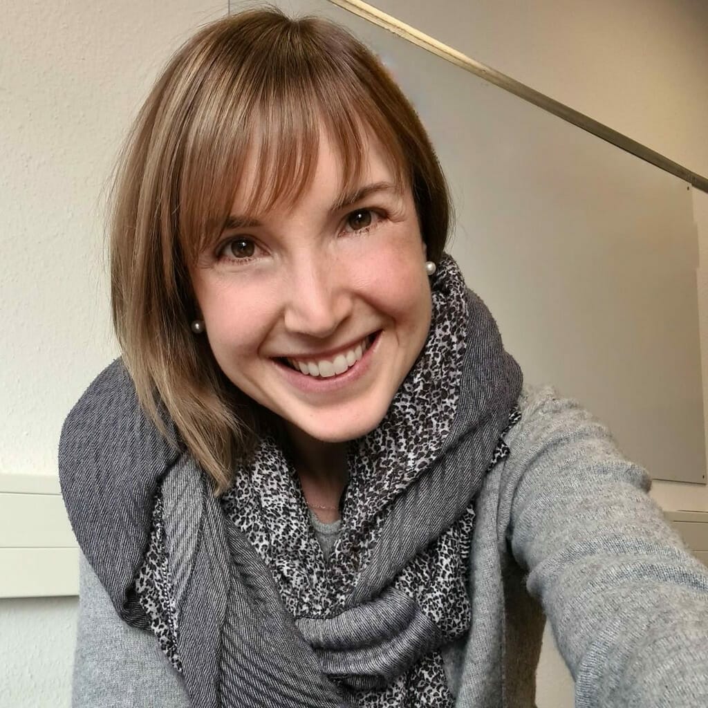 A person with short hair and a gray scarf smiles while looking directly at the camera. They are indoors with a light-colored wall and a framed board in the background, perhaps getting ready to attend a customer advisory board meeting.