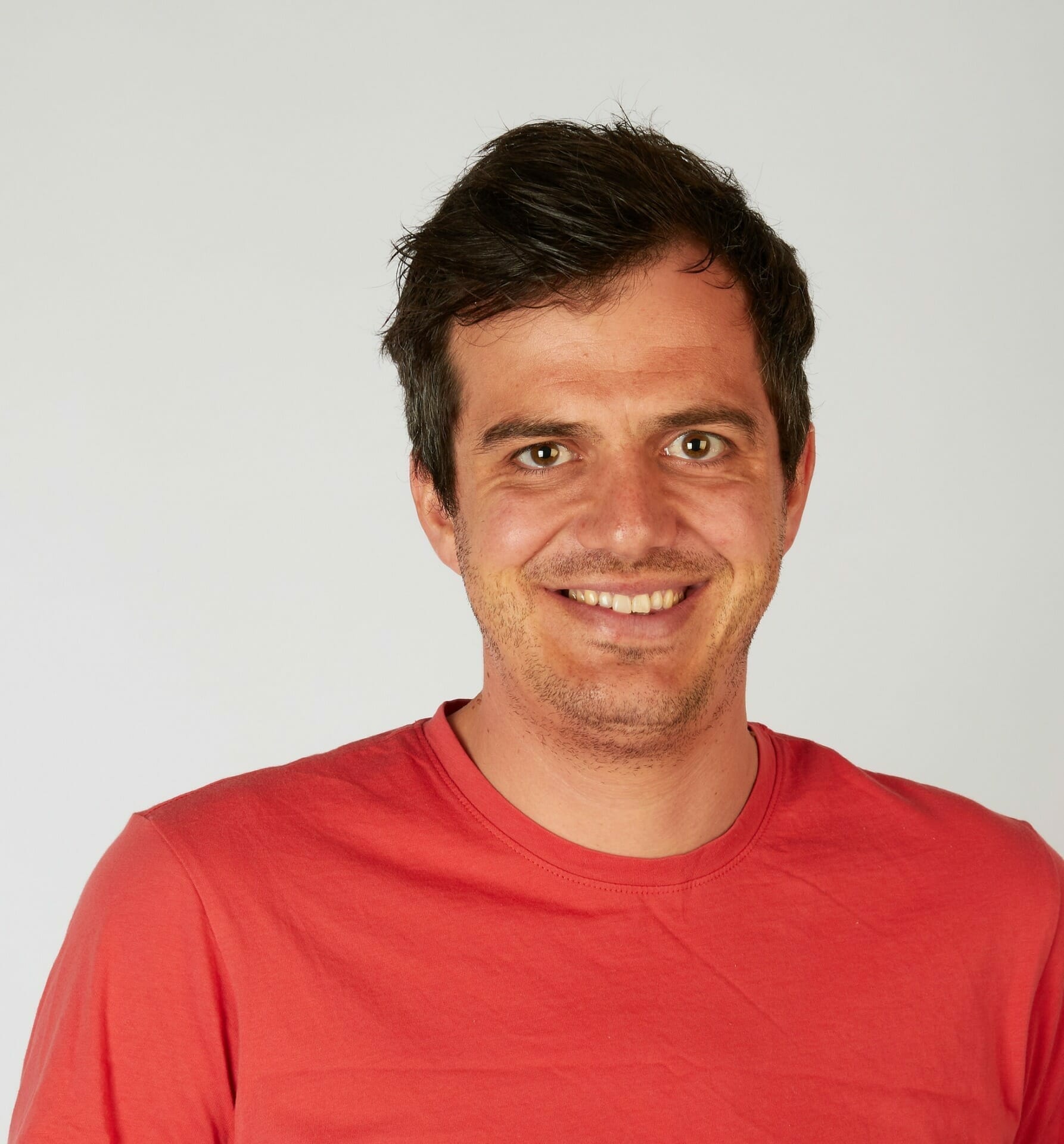 A person with short dark hair and a faint beard smiles at the camera, wearing a red T-shirt against a plain background, as if mid-discussion in a customer advisory board meeting.
