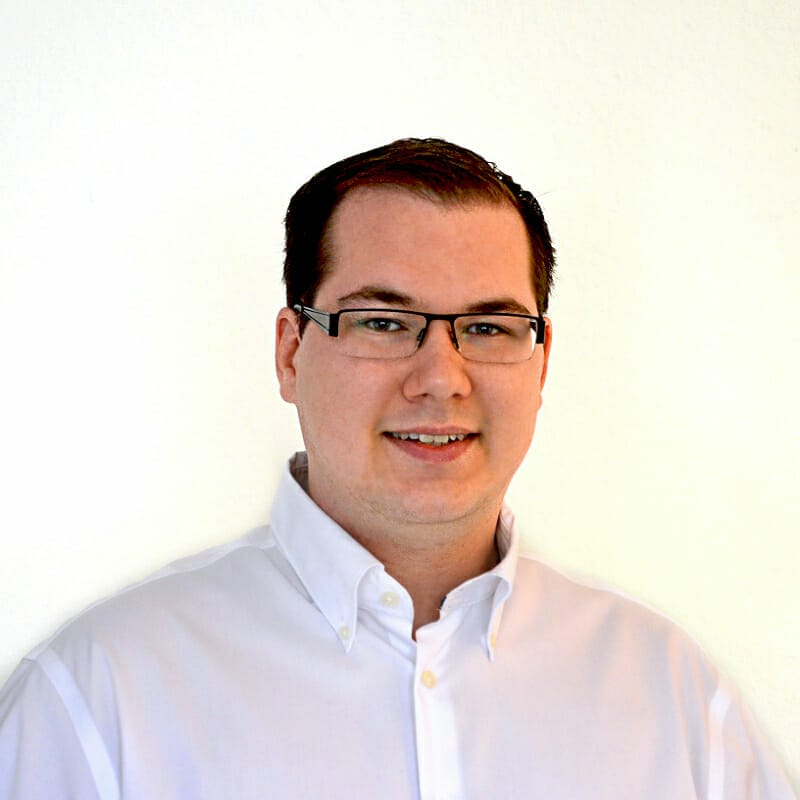 A man with short dark hair, wearing glasses and a white button-up shirt, stands in front of a plain white background, ready to go on air.