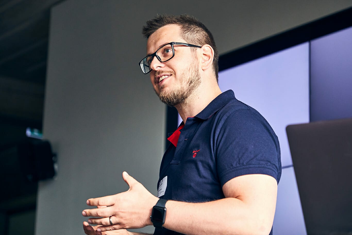 A man with glasses and a beard is giving a presentation, wearing a navy blue shirt and a smartwatch. He is gesturing with his hands and speaking in front of a screen, possibly sharing DIY tips.