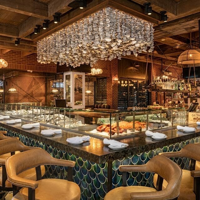 A modern seafood restaurant in Las Vegas with a central oyster bar, leather chairs, and a shell chandelier hanging from a wooden ceiling provides the perfect setting for culinary shoptalk.