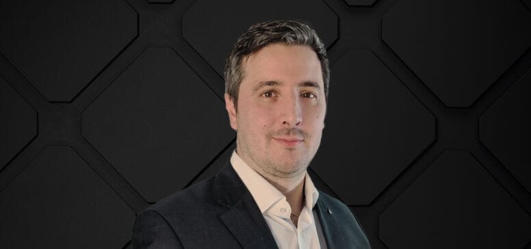 A man exuding leadership in a suit and light shirt stands confidently against a dark geometric background.